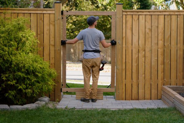 "A man wearing work gloves and a tool belt stands on a backyard patio while installing or aligning a wooden fence gate, with a green shrub on the left and a raised garden bed on the right