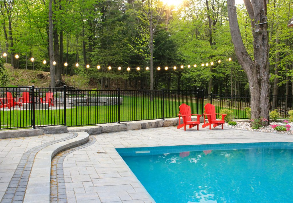 an iron fence or metal fence around a pool in a backyard with trees and a flower pot with plants and a sunset with string lights and chairs
