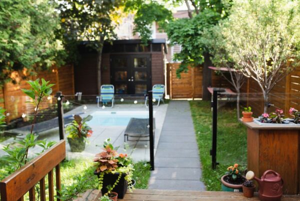 A cozy and newly renovated small backyard displaying a charming wooden deck adorned with potted plants. A neat concrete pathway runs through the center, bordered on the left by a clear blue swimming pool and on the right by a fresh wooden privacy fence. Overhead, string lights add a warm and inviting glow, enhancing the intimate and serene atmosphere of this compact outdoor sanctuary.