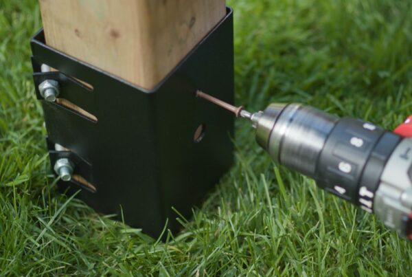 A close-up photo showing the process of securing a black metal post base into the ground with a drill. The image captures a drill bit inserted into a guide hole of the post base, which is attached to a wooden post. The background is a lush green lawn, emphasizing the outdoor setting of the construction project.