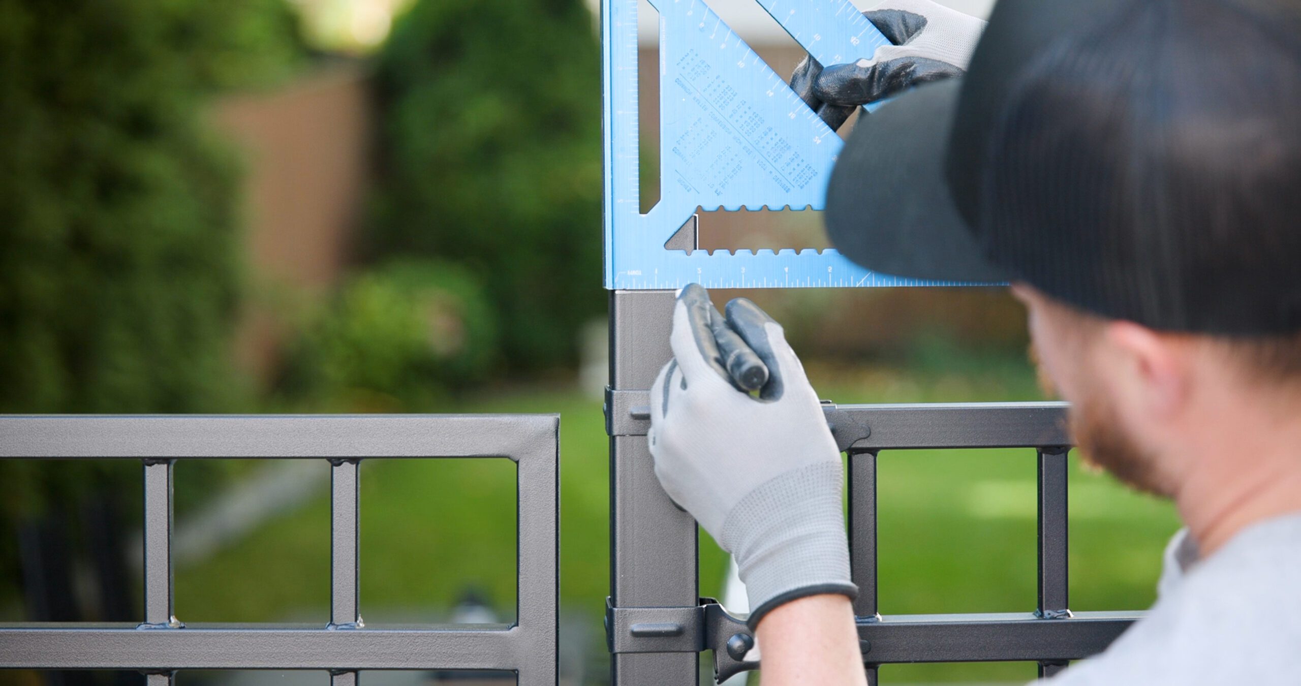 A person wearing gloves and a black cap using a blue carpenter square to mark precise measurements on a metal fence or gate post during installation, with a garden background.