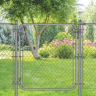 Dark gray chain-link fence with a matching walk-through gate installed in a lush backyard, framed by a willow tree, evergreen shrubs, and blooming pink flowers on a green lawn.
