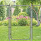 Gray chain-link fence with a matching walk-through gate installed in a backyard, surrounded by a willow tree, evergreen shrubs, and blooming pink flowers on a green lawn.