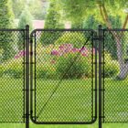 Black chain link gate installed in a lush backyard setting with green trees, shrubs, and pink flowering plants in the background.