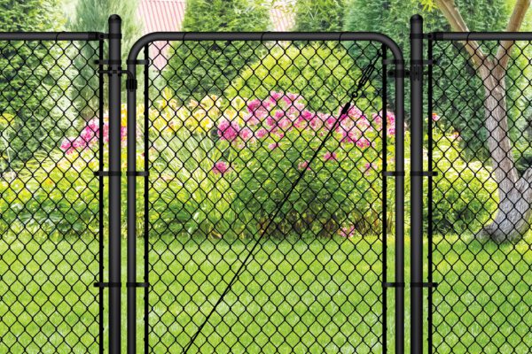 Black chain link gate installed in a lush backyard setting with a tree, evergreen shrubs, and pink flowering plants in the background.