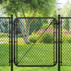 Black chain link gate installed in a lush backyard setting with a tree, evergreen shrubs, and pink flowering plants in the background.