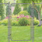Galvanized silver chain link gate installed in a green backyard setting with a tree, evergreen shrubs, and pink flowering plants in the background